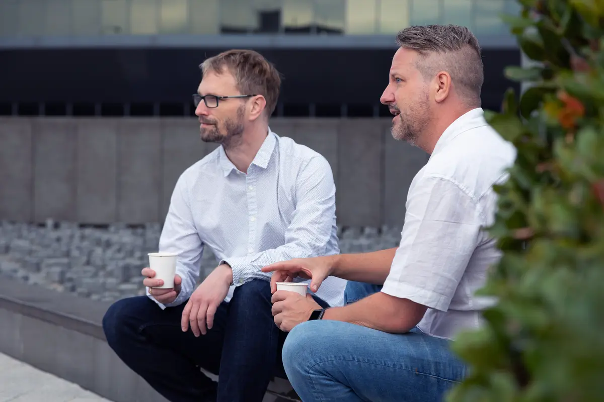 Two men sitting outside holding coffee cups and talking