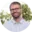 A bearded man wearing glasses and a dotted shirt smiles outdoors.