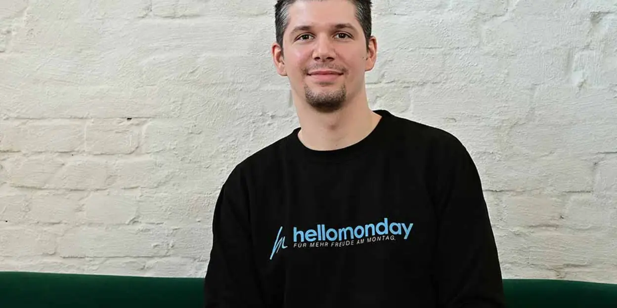 Persona con suéter negro con texto 'hellomonday' sentado contra una pared de ladrillo