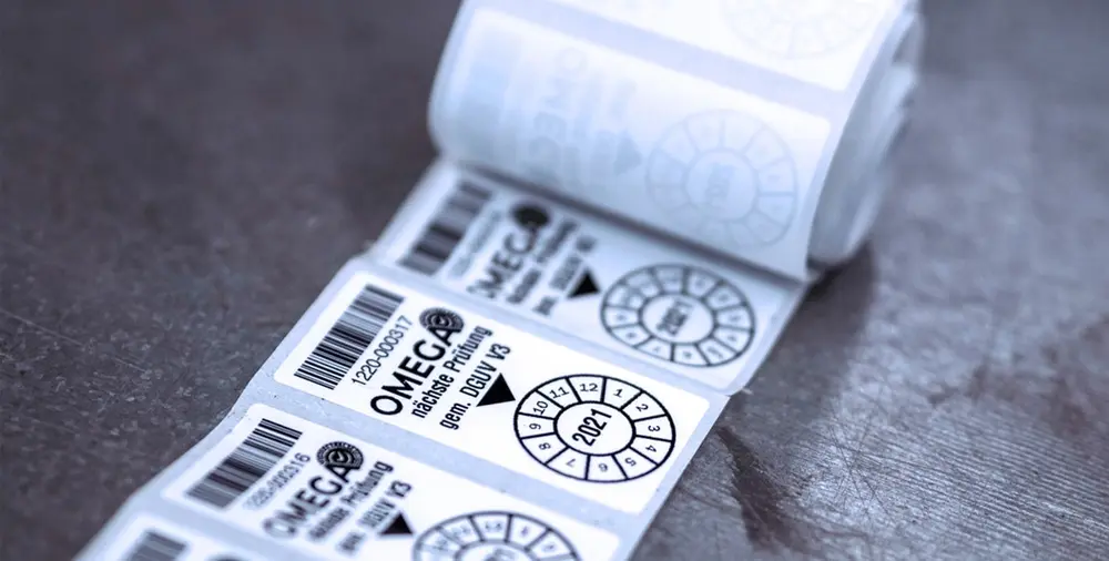Close-up of a roll of inspection stickers with OMEGA branding on a grey surface.