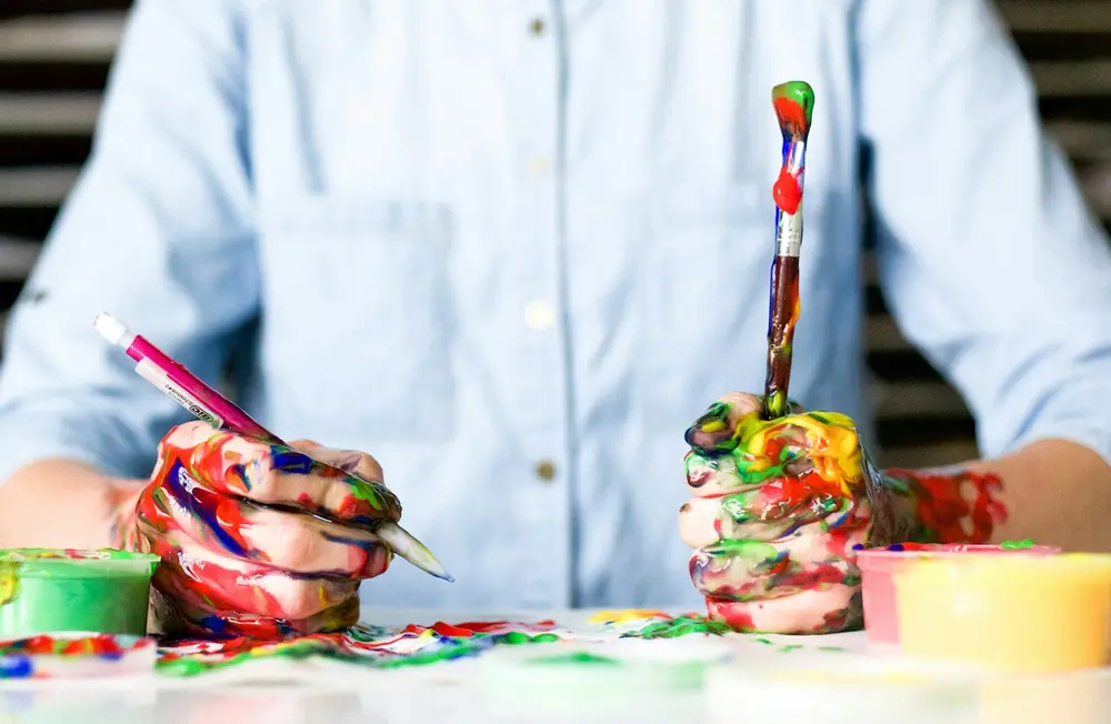 Hands covered in paint holding a brush and pen