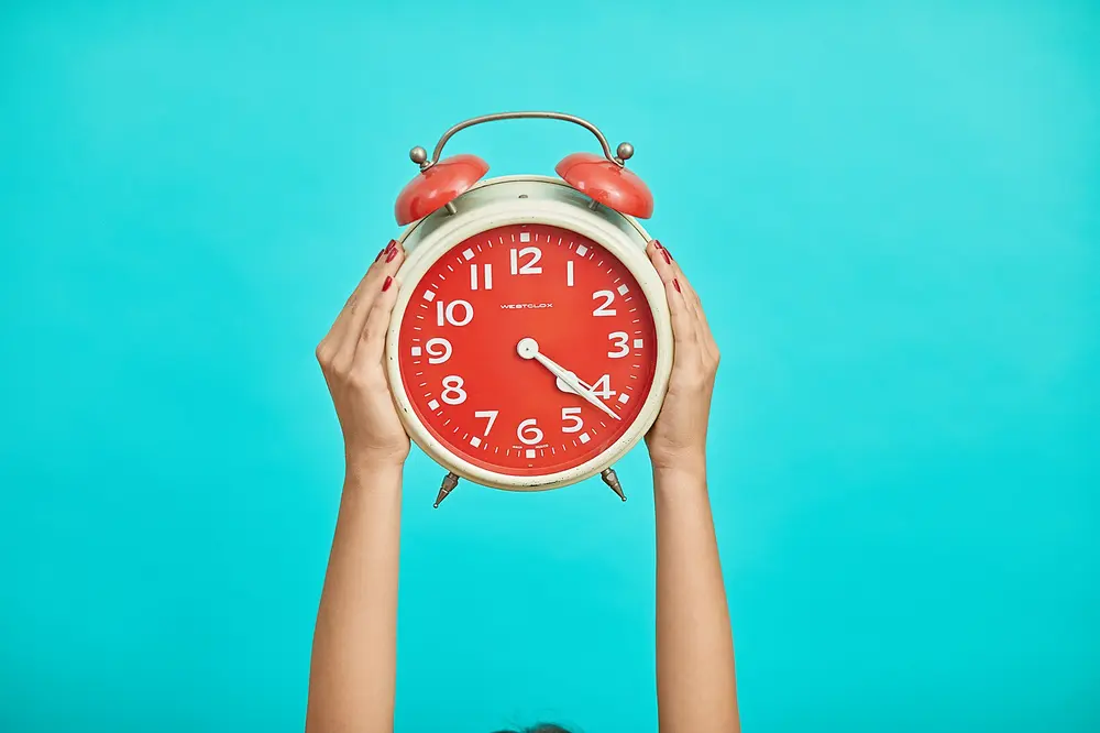 Hands holding a red alarm clock against a bright blue background